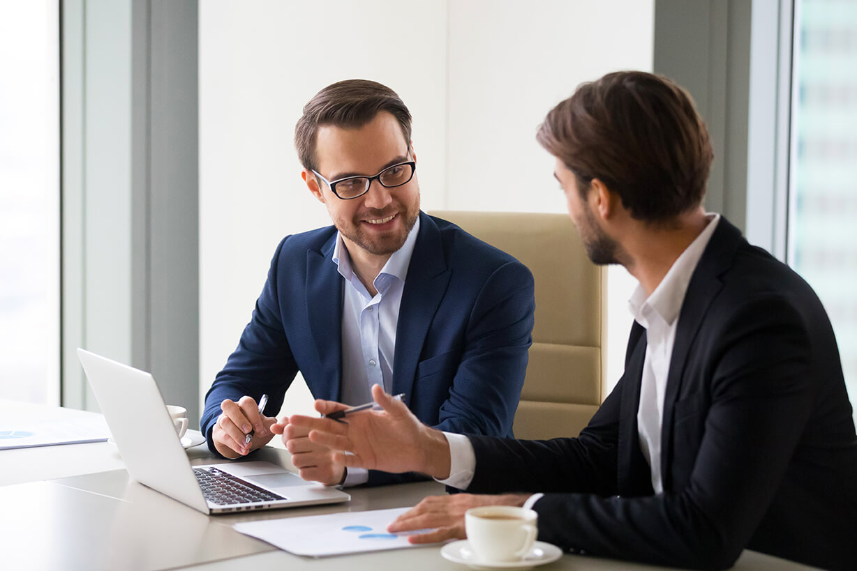 Two businessmen sat at an office table, talking in front of a laptop. 