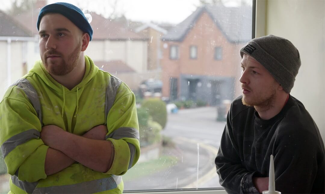 Two men wearing woolen caps, standing by a suburban window