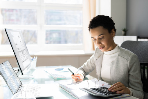 A young female adult with short cropped black hair is sat at a desk beside a window, managing her business accounts. She wears an open front smart grey jacket over a high-neck cream top. She holds a pen in one hand, the other poised at a large calculator. She is focused on an open, ring-bound file of work. A laptop and PC are also on the desk, displaying information.