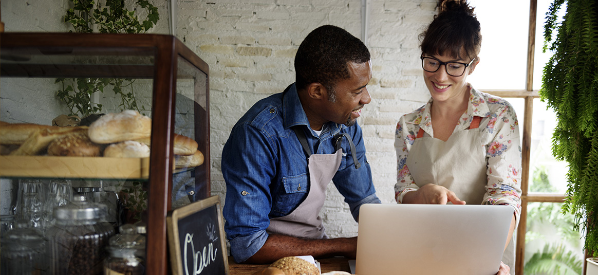 Photo of two people in aprons talking in front of a laptop on a café counter.
