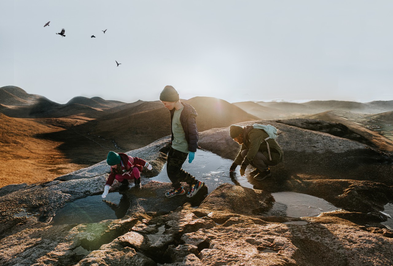 A man kneels by a frozen puddle and pokes at the ice. His young daughter does the same. A boy walks over the uneven, rocky terrain of a mountain peak. The family enjoys this outdoor activity together. The sun is low in the sky. Depicts camping, hiking, exploring, tourism etc.